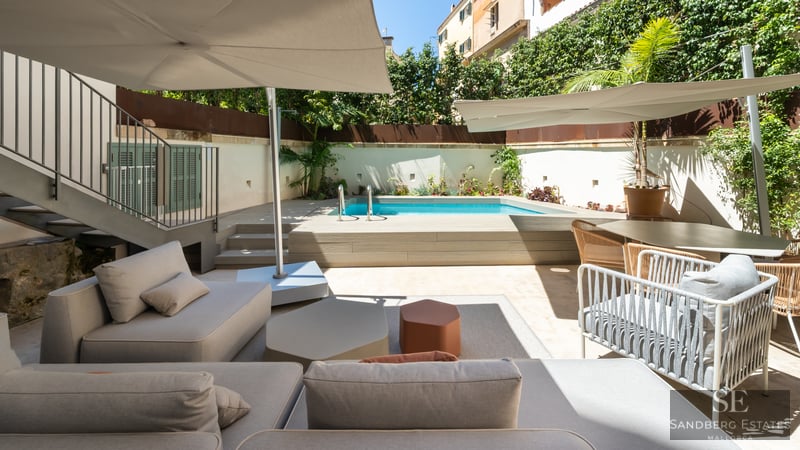 Modern terrace featuring a turquoise pool, wooden deck, grey sofas, and sun umbrellas under a clear sky.
