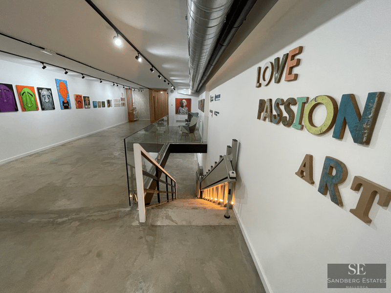 Galería de arte moderna con pinturas coloridas, suelos de cemento, una escalera de cristal y un elevador para sillas de ruedas.