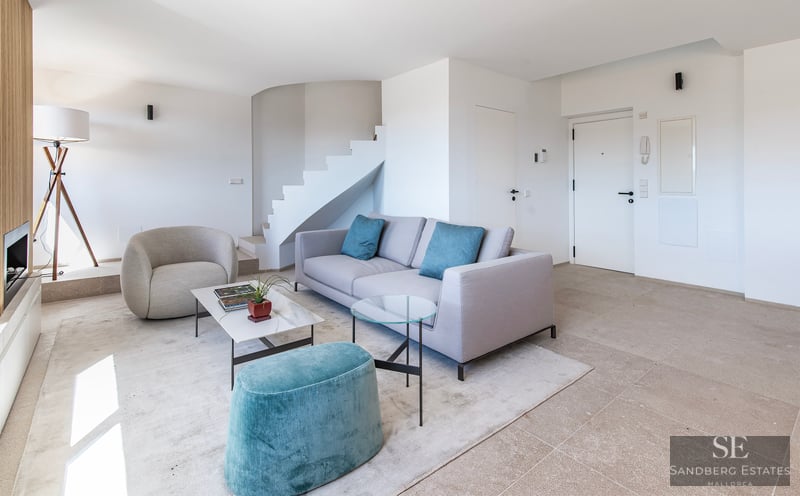 Sala de estar minimalista con sofá gris, otomana turquesa, escalera blanca y mesa de mármol.