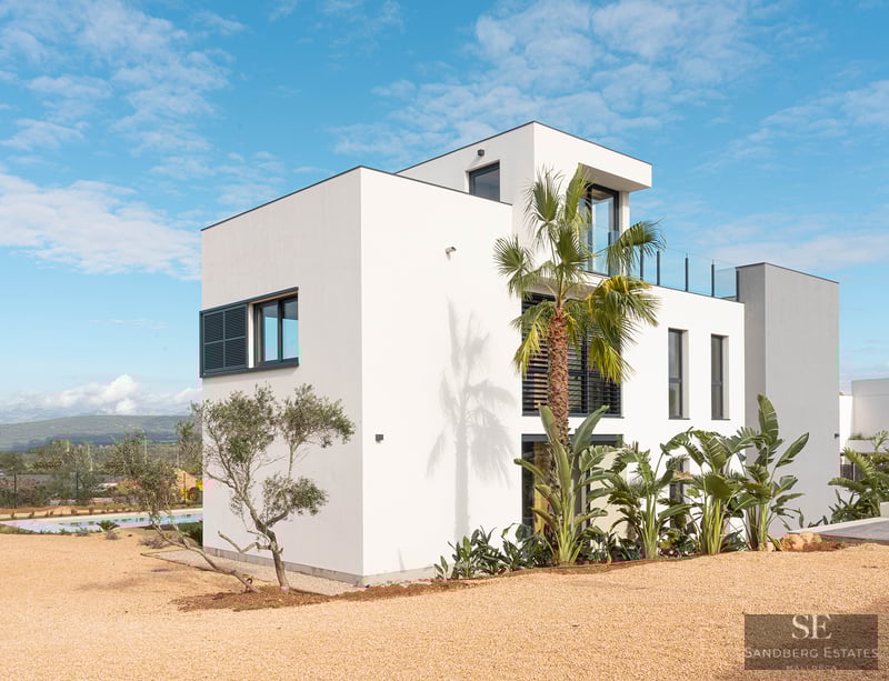 Exterior of a three-level white villa with modern architecture, palm trees, and a gravel yard under a blue sky.