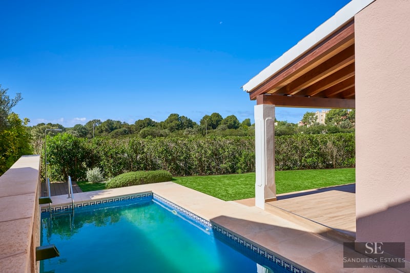 A turquoise swimming pool next to a green lawn and a covered terrace with wooden beams under a clear blue sky.