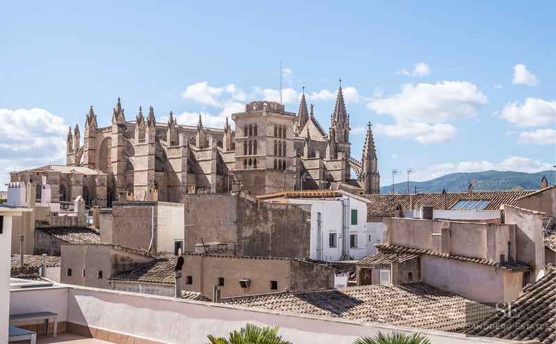 Une grande cathédrale gothique surplombe les toits en terre cuite d'une ville méditerranéenne sous un ciel bleu.