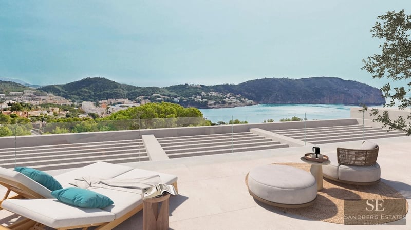 Modern stone terrace with sun loungers and designer furniture overlooking a Mediterranean bay and green hills.