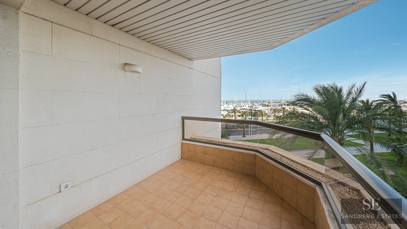 Covered terrace with glass railing overlooking a harbor with yachts and palm trees under a clear blue sky.