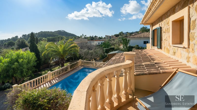 View from a stone balcony overlooking a blue swimming pool, lush gardens, and rolling hills under a clear blue sky.