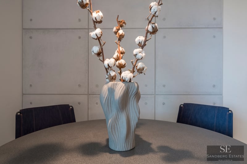 Jarrón gris texturizado con ramas de algodón sobre una mesa oscura frente a una pared de paneles de hormigón.