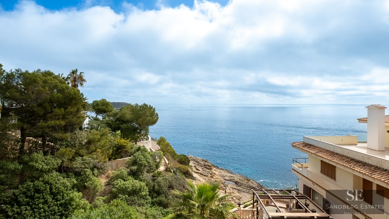 Elevated view of a rocky coastline with deep blue sea, green pine trees, and a Mediterranean villa.