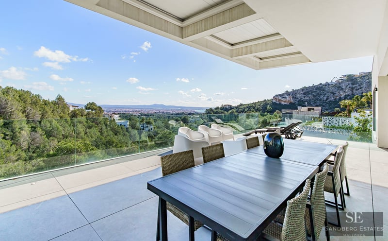 Modern terrace with a dining table and glass railings overlooking a lush green forest and distant city.
