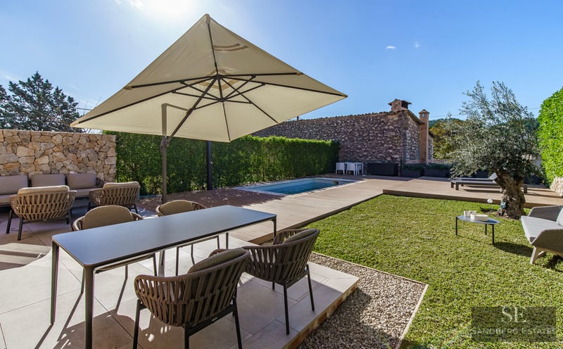 Sunlit terrace with modern dining set, large umbrella, small swimming pool, and lawn area with an olive tree.