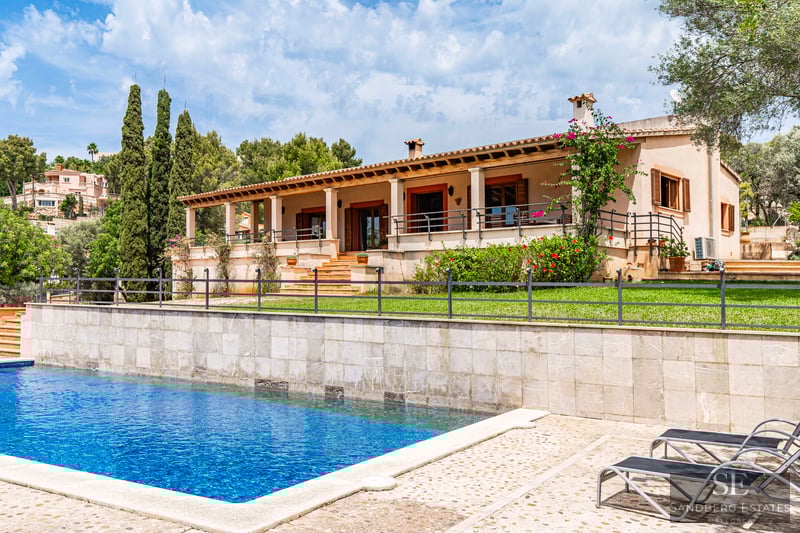Gran piscina azul y tumbonas frente a una villa mediterránea con muro de piedra, jardín verde y terraza cubierta.