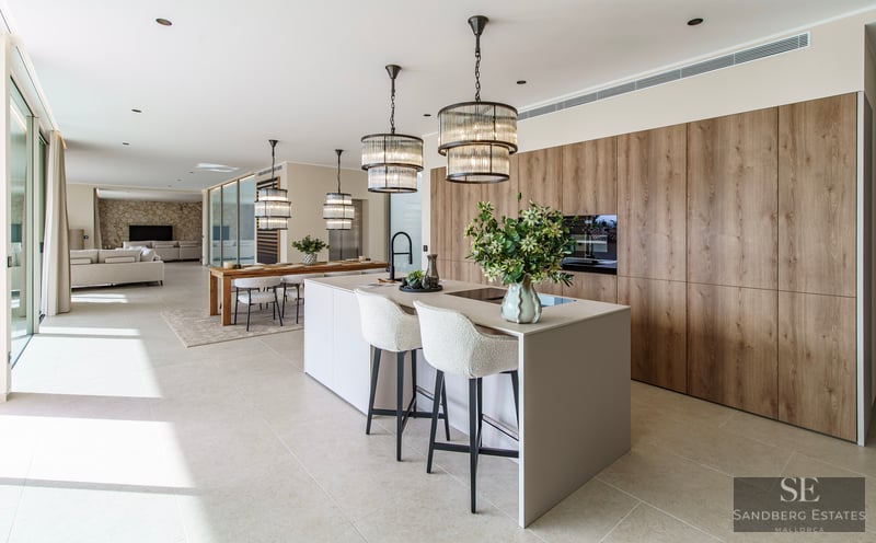 Modern kitchen with a large island, white stools, designer pendant lights, and wood cabinetry in an open-plan space.