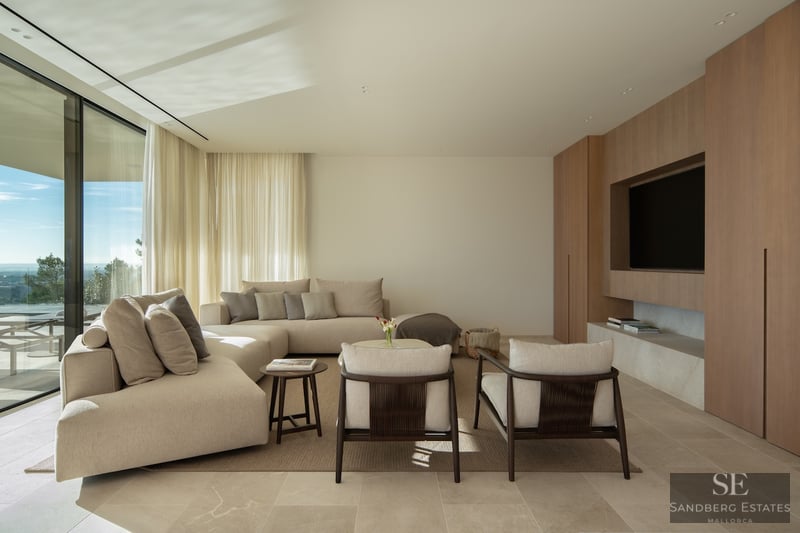 Modern living room with a beige sectional sofa, wood paneled TV wall, and floor-to-ceiling windows with a scenic view.