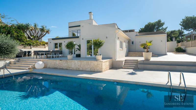 Piscine turquoise devant une villa blanche moderne avec terrasse en pierre sous un ciel bleu clair.
