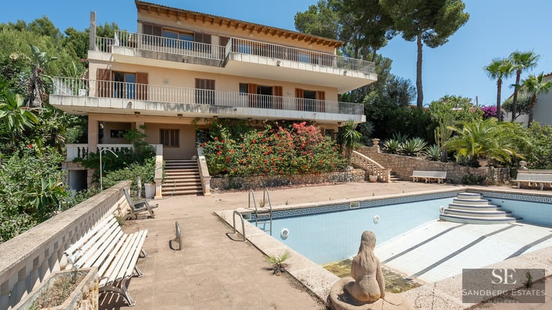 Multi-level Mediterranean villa featuring large terraces and a large empty swimming pool surrounded by lush greenery.