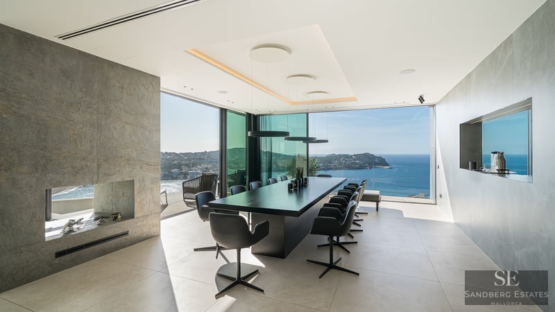 A modern dining room with a long black table, stone fireplace, and floor-to-ceiling windows overlooking the ocean.