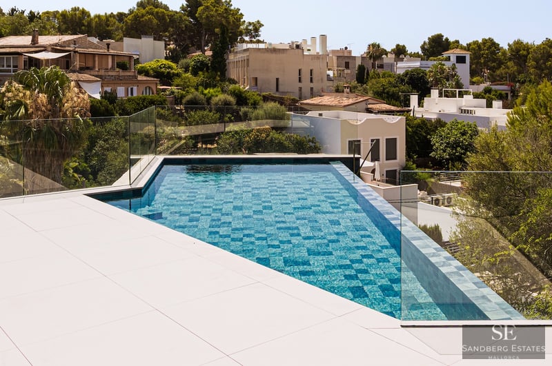 High-angle view of a triangular turquoise swimming pool with glass railings and a white tiled deck overlooking a town.