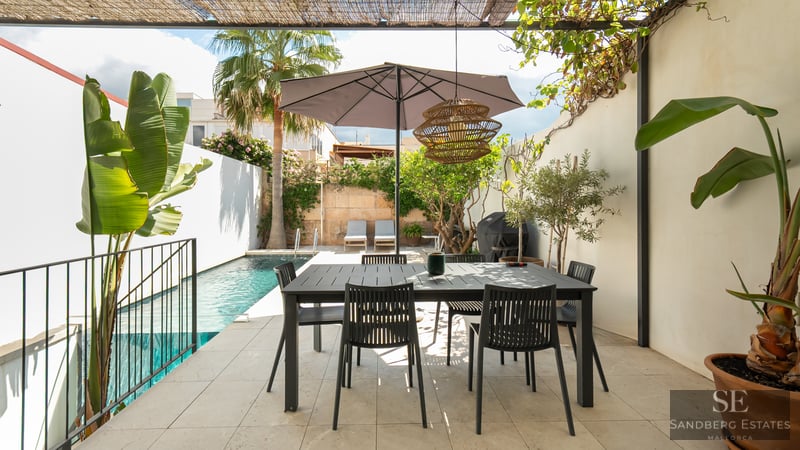 Terraza moderna con mesa de comedor negra bajo sombrilla, junto a una piscina turquesa y palmeras.