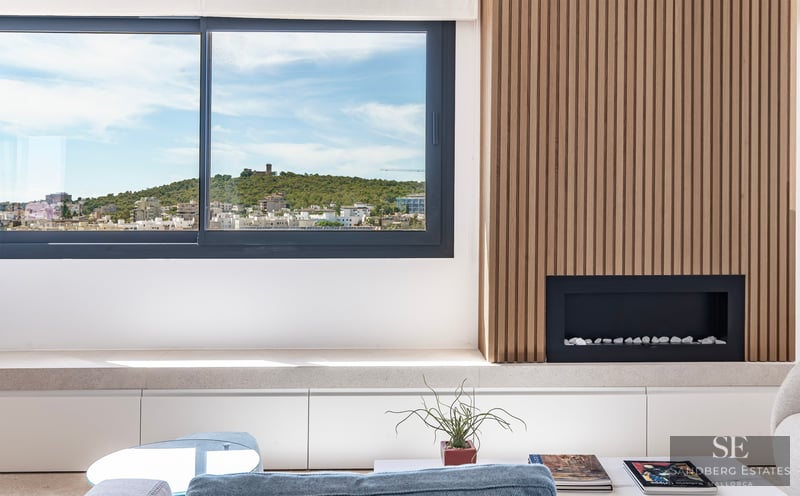Salón moderno con pared de madera, chimenea integrada y ventanal con vistas a una colina con un castillo.