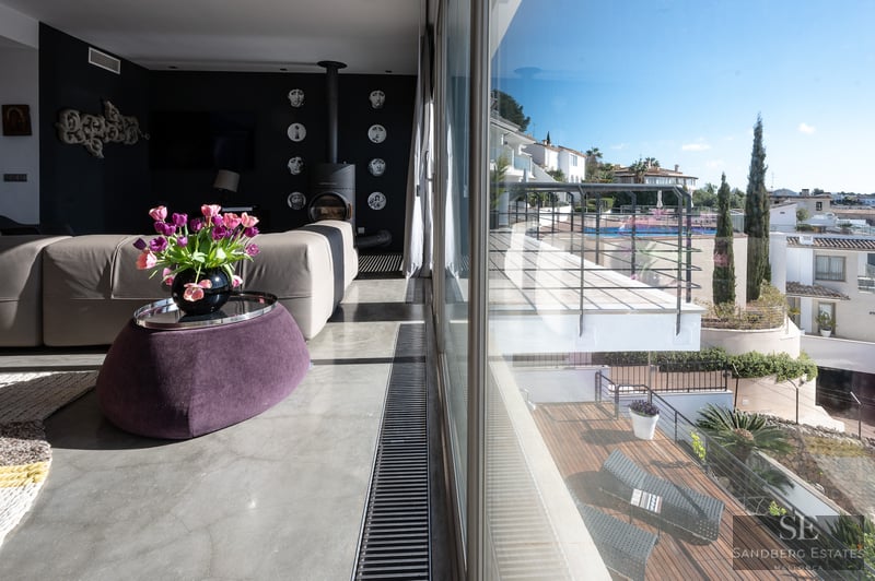 Salón moderno con suelo de hormigón, sofá beige, puf morado y vistas a una terraza soleada a través de grandes cristales.