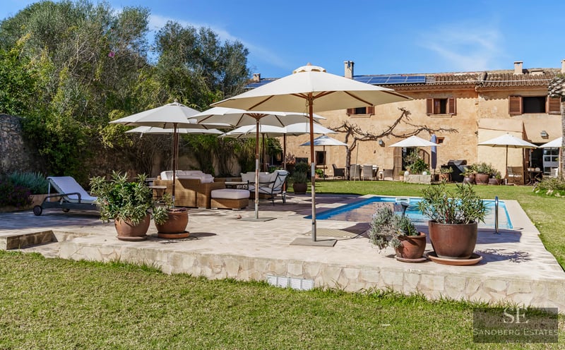 Large blue swimming pool surrounded by a stone deck with white umbrellas and wicker lounge furniture in a sunny garden.