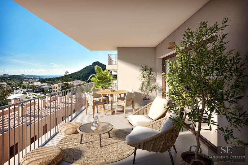 Modern balcony with woven furniture, dining table, and plants overlooking a scenic coastal town and green hills.