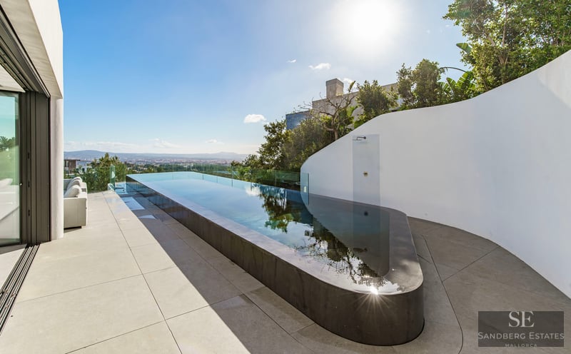 A sleek, long infinity pool on a minimalist white terrace overlooking a distant city under a bright blue sky.