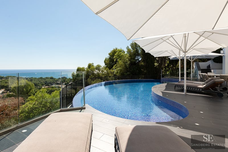 A curved blue swimming pool on a modern stone terrace with white umbrellas and sunbeds overlooking the sea.