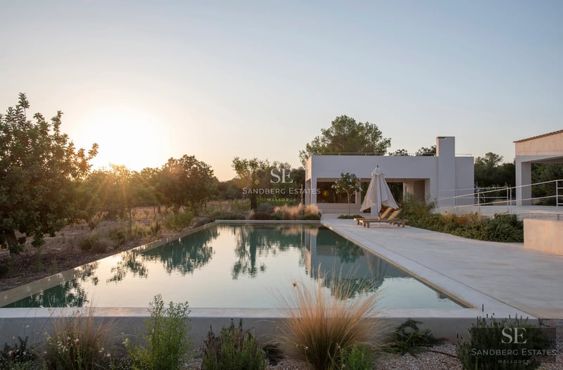 Vista exterior de villa moderna con piscina infinita, terraza, y jardín. Diseño minimalista, iluminación natural y vistas panorámicas.