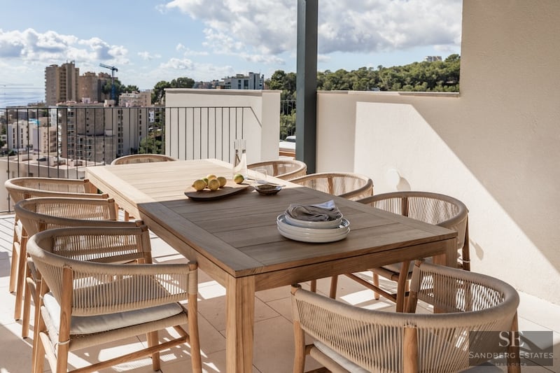 Luxury Terrace Dining with Sea and City Views Modern outdoor dining area on a terrace featuring a wooden table, woven chairs, and a panoramic sea view.