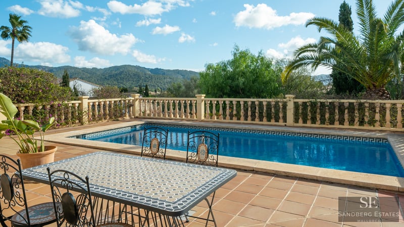 Rectangular swimming pool and mosaic dining table on a terracotta terrace with mountain views and palm trees.