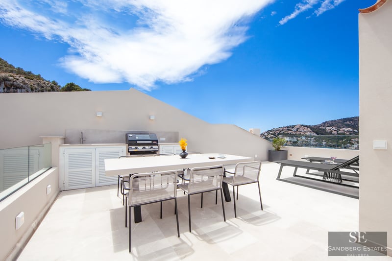 Modern rooftop terrace featuring an outdoor kitchen, dining table, and sun loungers with panoramic coastal views.