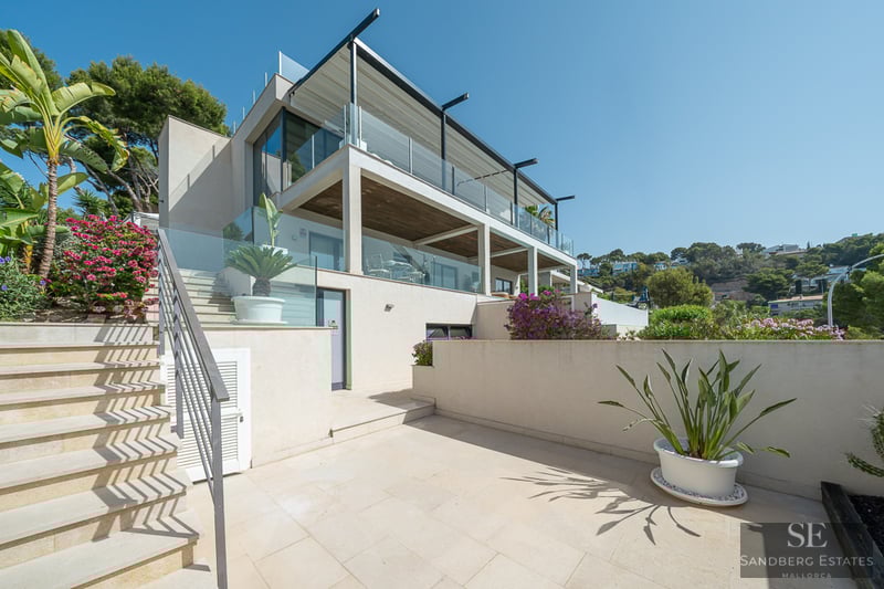 Mehrstöckige moderne Villa mit beigefarbenen Steintreppen, Glasgeländern und Bougainvillea unter klarem blauem Himmel.