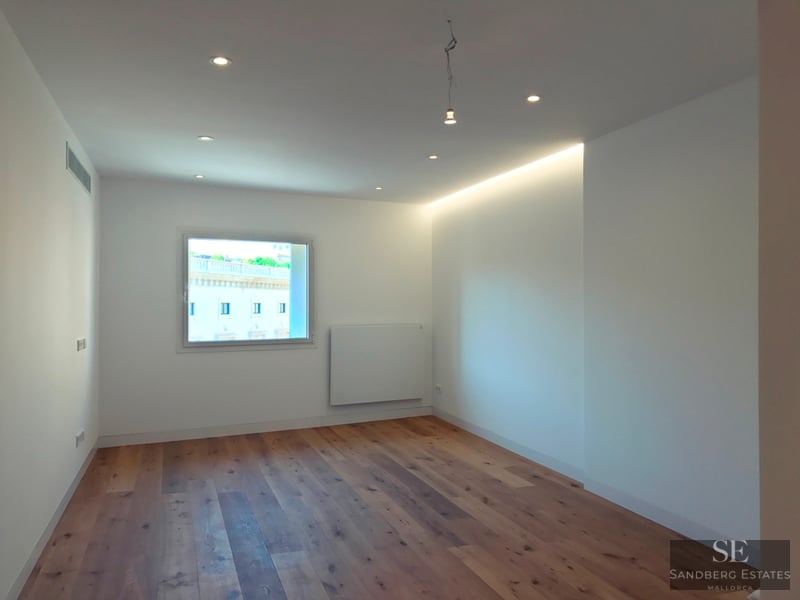 Empty white room with light oak wood floors, recessed ceiling lights, and a window showing a classical building outside.