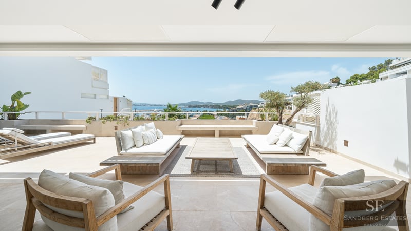 Großzügige weiße Terrasse mit Holzsofas und Sesseln mit Blick auf das blaue Meer und die Küste unter hellem Himmel.