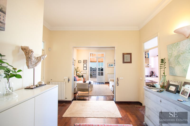 Flur mit Holzböden, der in ein sonniges Wohnzimmer mit Sofas und Zugang zu einem Außenbalkon führt.