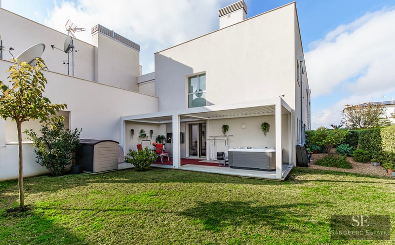 A modern white building with a green lawn and a covered terrace area featuring a hot tub and dining set.