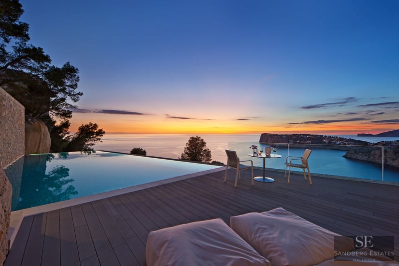 Infinitypool på trädäck med utsikt över havet vid solnedgången, utemöbler och glasräcke.