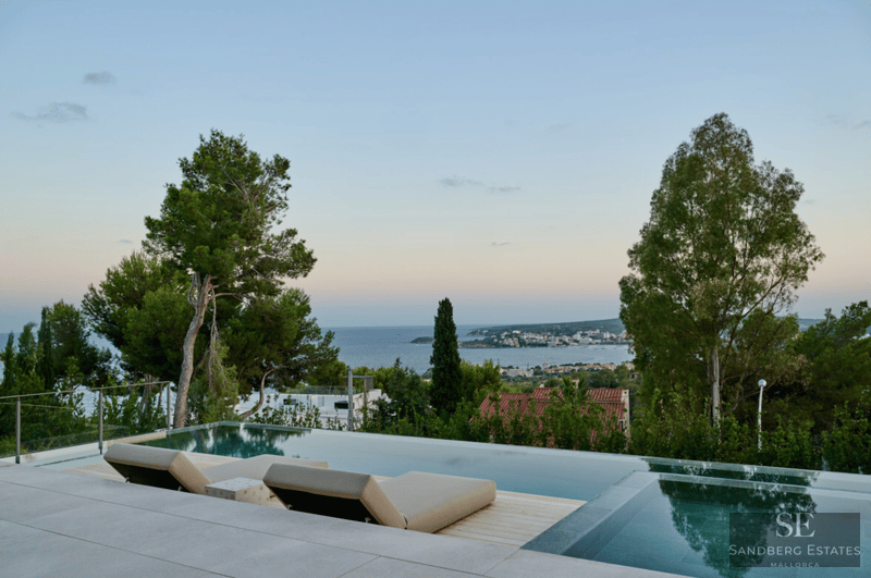 Piscina infinita moderna con dos tumbonas beige frente al mar azul y una ciudad costera al atardecer.