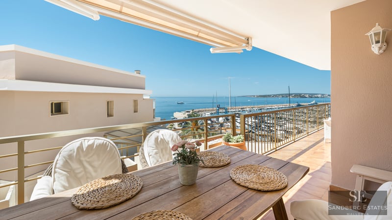 Balcony with wooden dining table and white chairs overlooking a marina and blue sea under a clear sky.