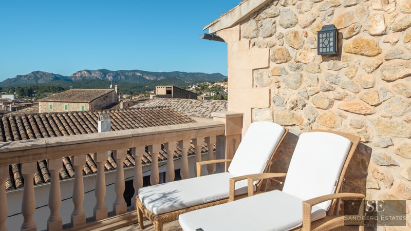 Dos tumbonas blancas en una terraza de piedra con vistas a un pueblo mediterráneo y montañas.