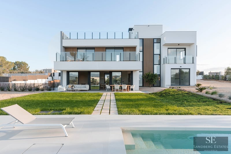 Modern white two-story villa with a turquoise swimming pool, sun lounger, and a green lawn under a clear sky.