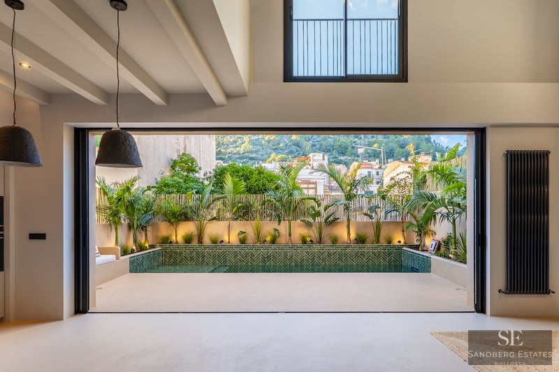 Vue intérieure par une grande baie vitrée vers une piscine privée entourée de plantes tropicales et carrelage vert.
