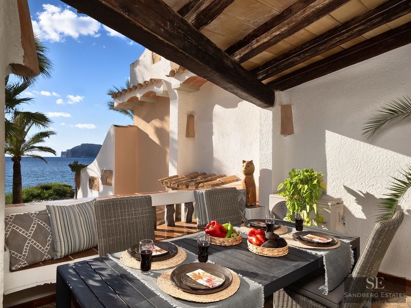 Terraza sombreada con mesa de comedor con vistas al mar azul, palmeras y acantilados bajo un cielo despejado.