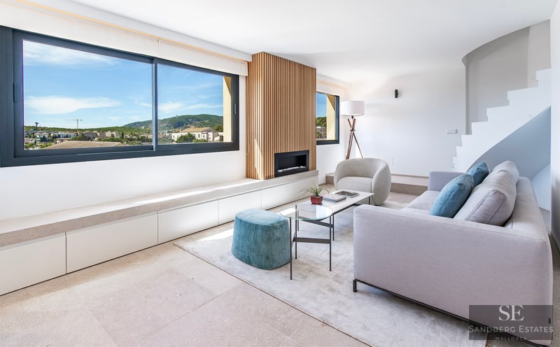 Salón luminoso con sofá gris, otomana turquesa, chimenea de listones de madera y gran ventanal con vistas.