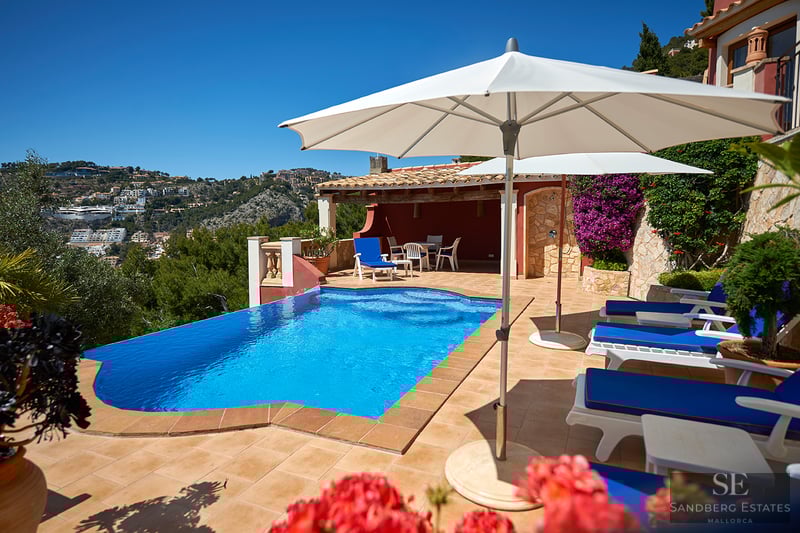 Privat poolterrass med blå solstolar och vita parasoll med utsikt över natursköna kullar.