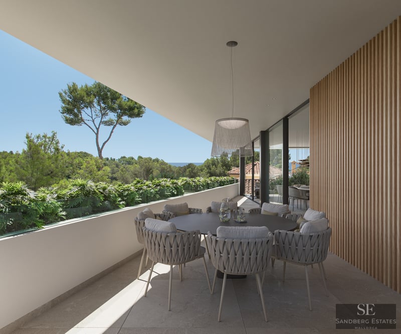 Modern terrace with a round dining table, woven chairs, wood-slatted wall, and a view of lush greenery and the sea.