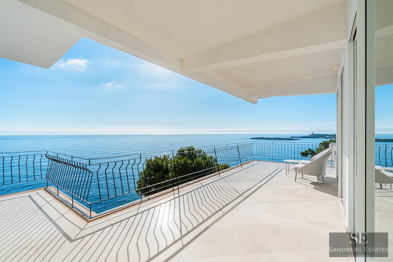 Bred modern terrass med stengolv och svart räcke med utsikt över ett lugnt blått hav under en klar himmel.