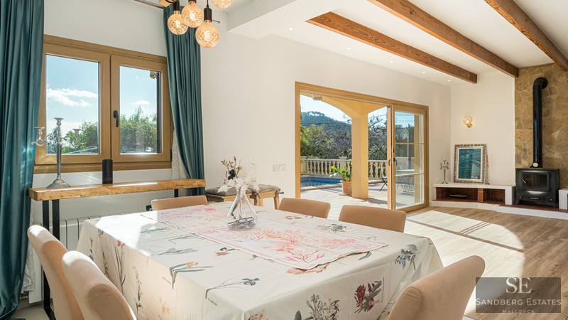 Bright dining room with wooden beams, stone fireplace, and glass doors leading to a terrace with a swimming pool.