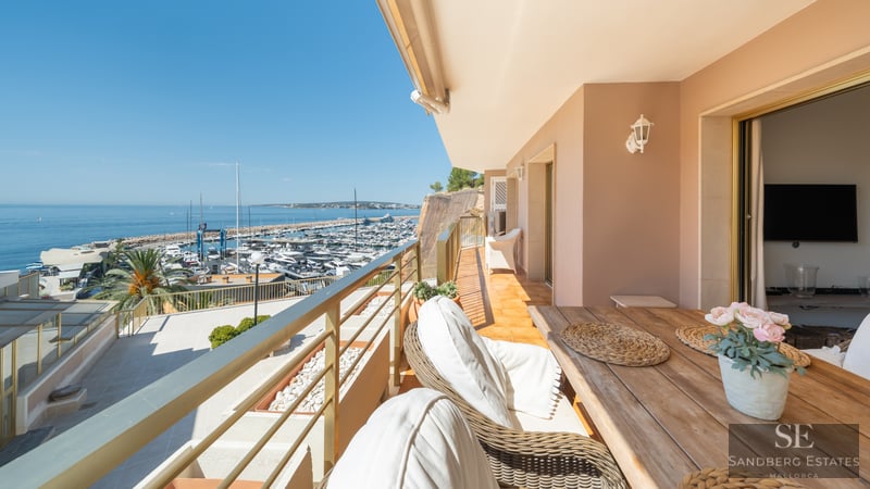 A sunlit balcony with a wooden dining table and wicker chairs overlooking a crowded marina and the blue ocean.