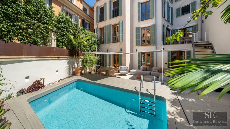 Light blue pool in a courtyard with wooden decking, outdoor sofas, and a building facade with green shutters.